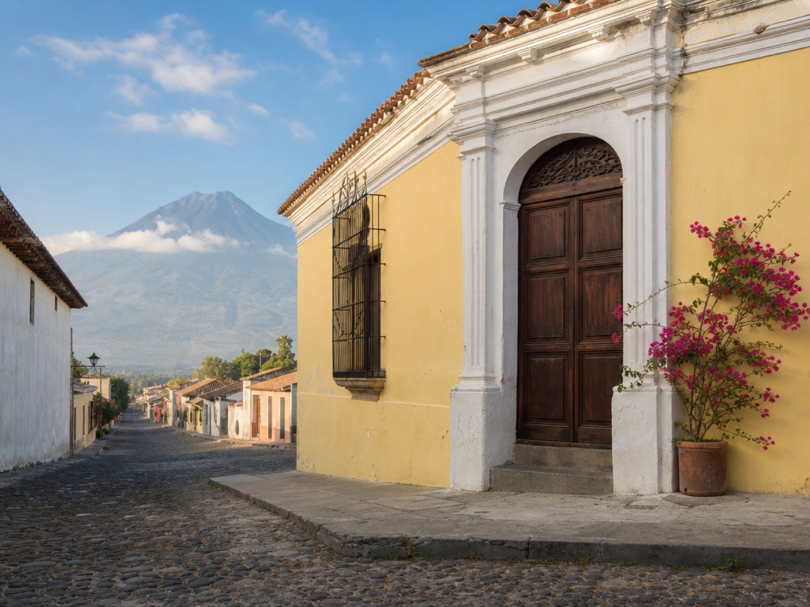 Colonial Antigua Guatemala street in bright morning light with a carved wooden door, bougainvillea and Volcán de Agua in the distance