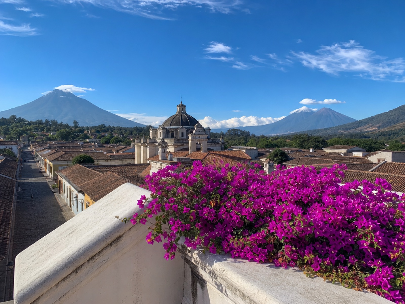 Rooftop view over colonial Antigua Guatemala on a clear dry-season morning with terracotta tile roofs, a whitewashed church dome, bougainvillea and Volcán de Agua under a blue sky