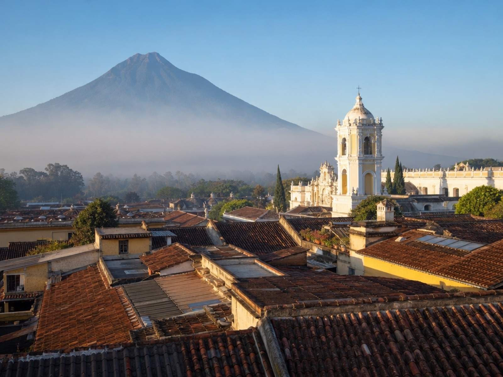 Elevated morning view over the terracotta rooftops and colonial church towers of Antigua Guatemala with Volcán de Agua rising behind the town in soft haze