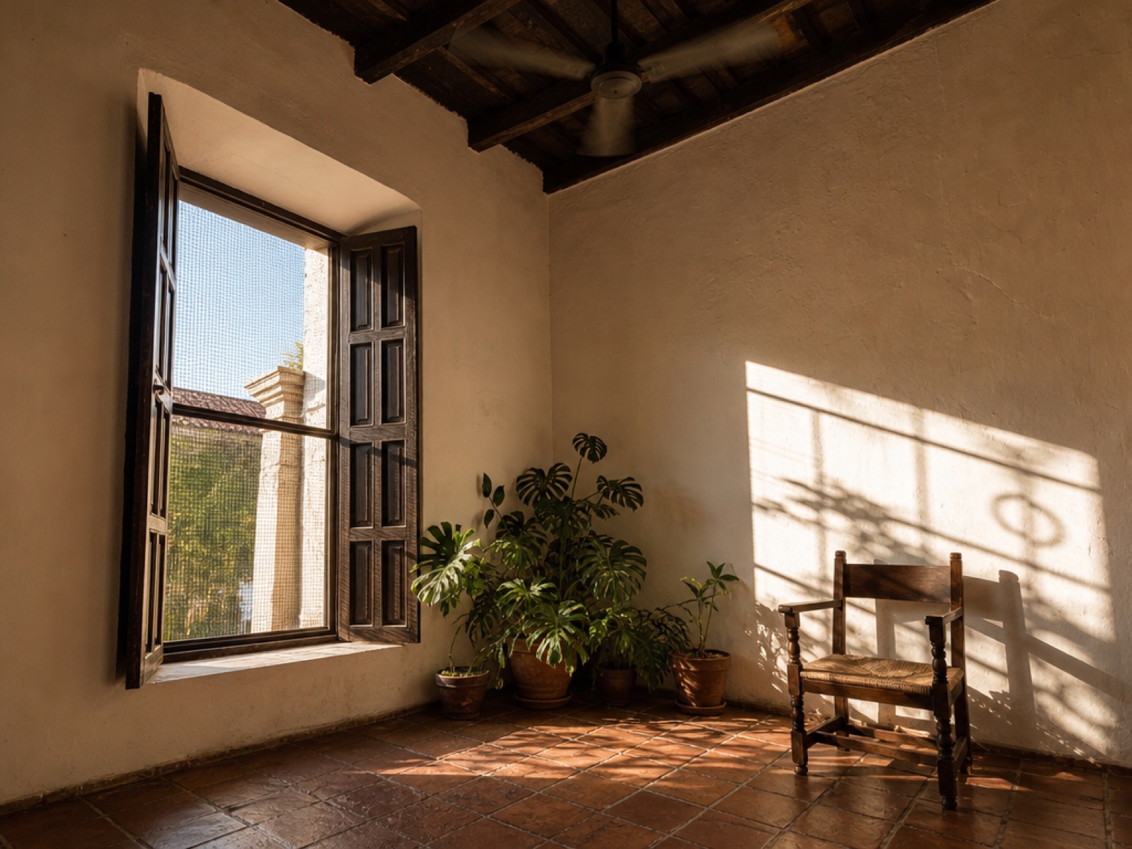 Sunlit corner of a colonial room in Antigua Guatemala with open wooden shutters and a mesh window screen, terracotta floor tiles, potted plants and a wooden chair under a beamed ceiling with a ceiling fan