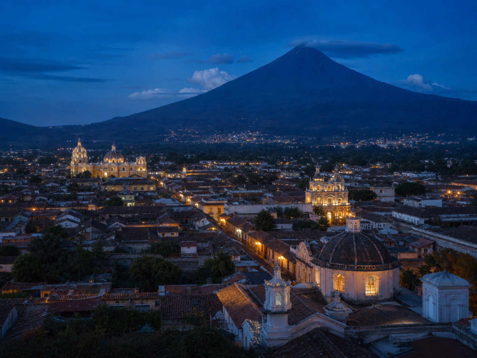 Illuminated colonial rooftops and church domes of Antigua Guatemala at blue hour beneath the dark cone of Volcán de Agua