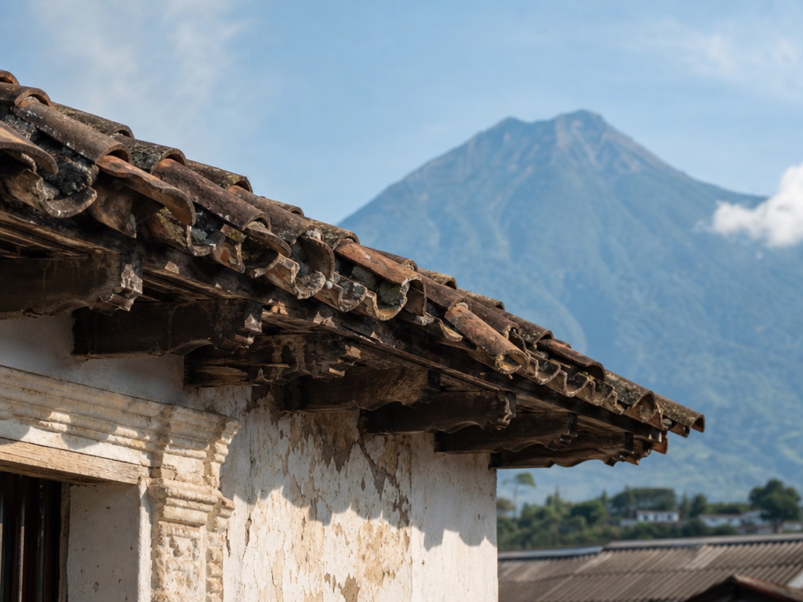 Weathered terracotta roof tiles and aged wooden eaves above a cracked white colonial wall in Antigua Guatemala, with Volcán de Agua rising in the background