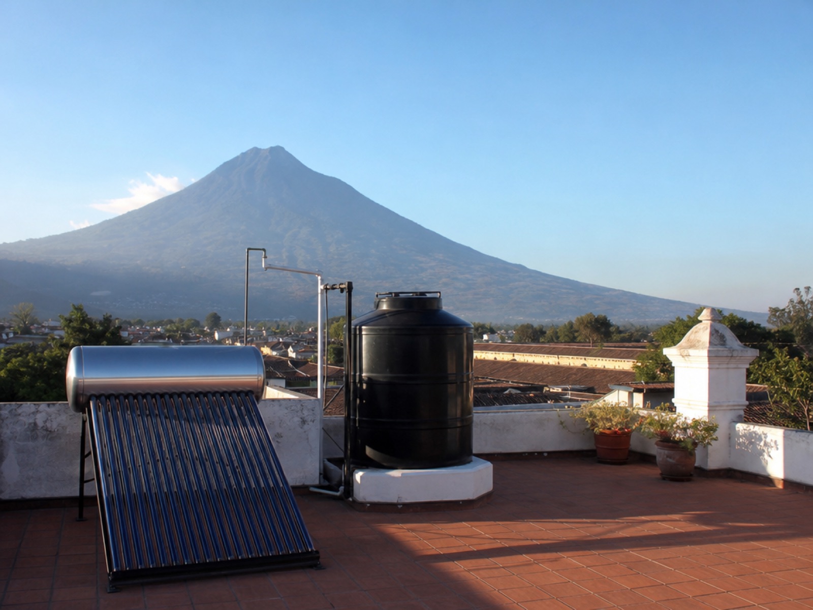 Rooftop of a colonial house in Antigua Guatemala with a solar water heater panel and a black water storage tank beside terracotta tiles, Volcán de Agua in the background