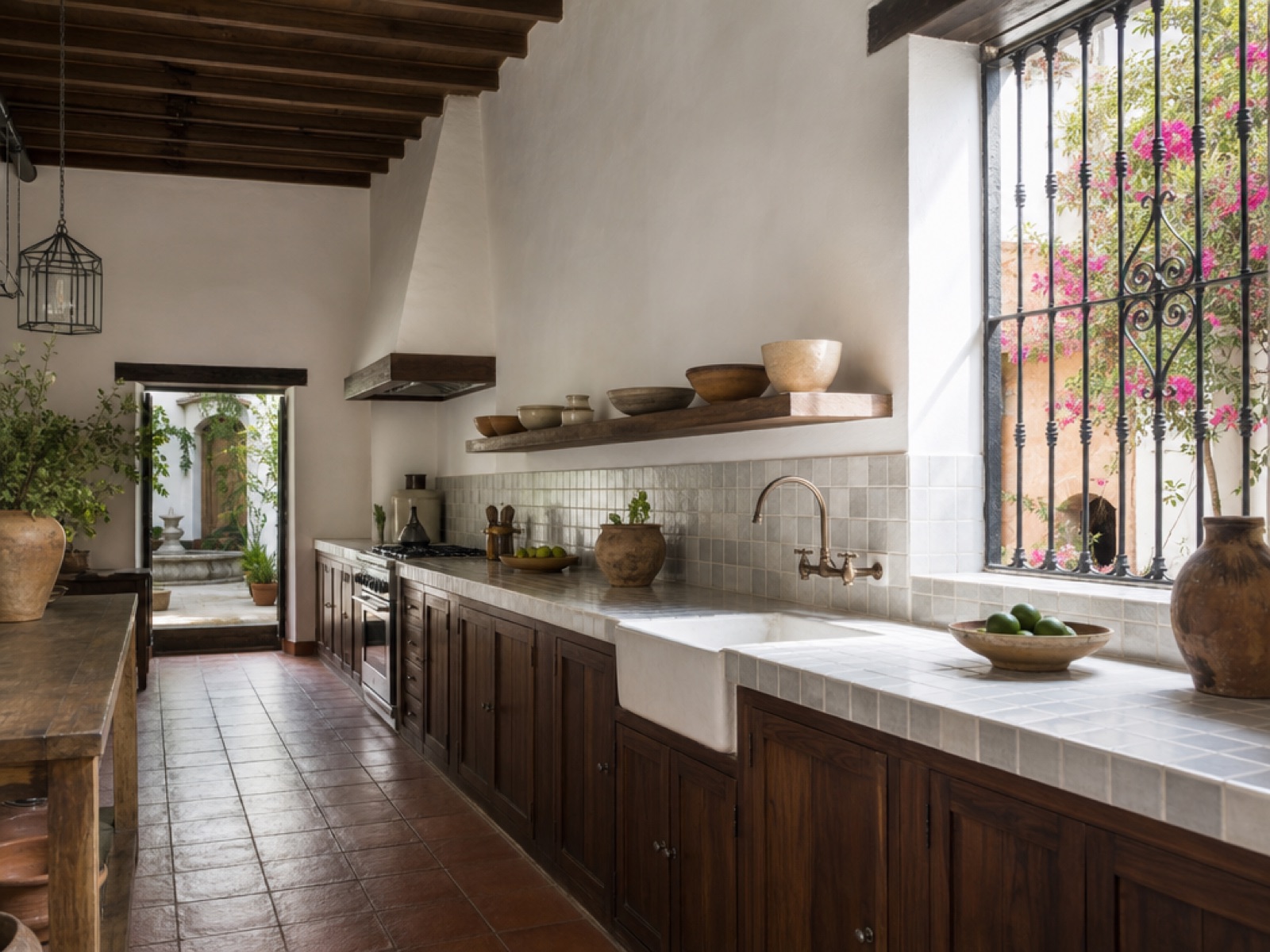 Renovated colonial kitchen in Antigua Guatemala with grey tiled countertop, dark wood cabinets and a farmhouse sink, timber ceiling beams, terracotta floor and a window with an iron grille open onto a bougainvillea courtyard