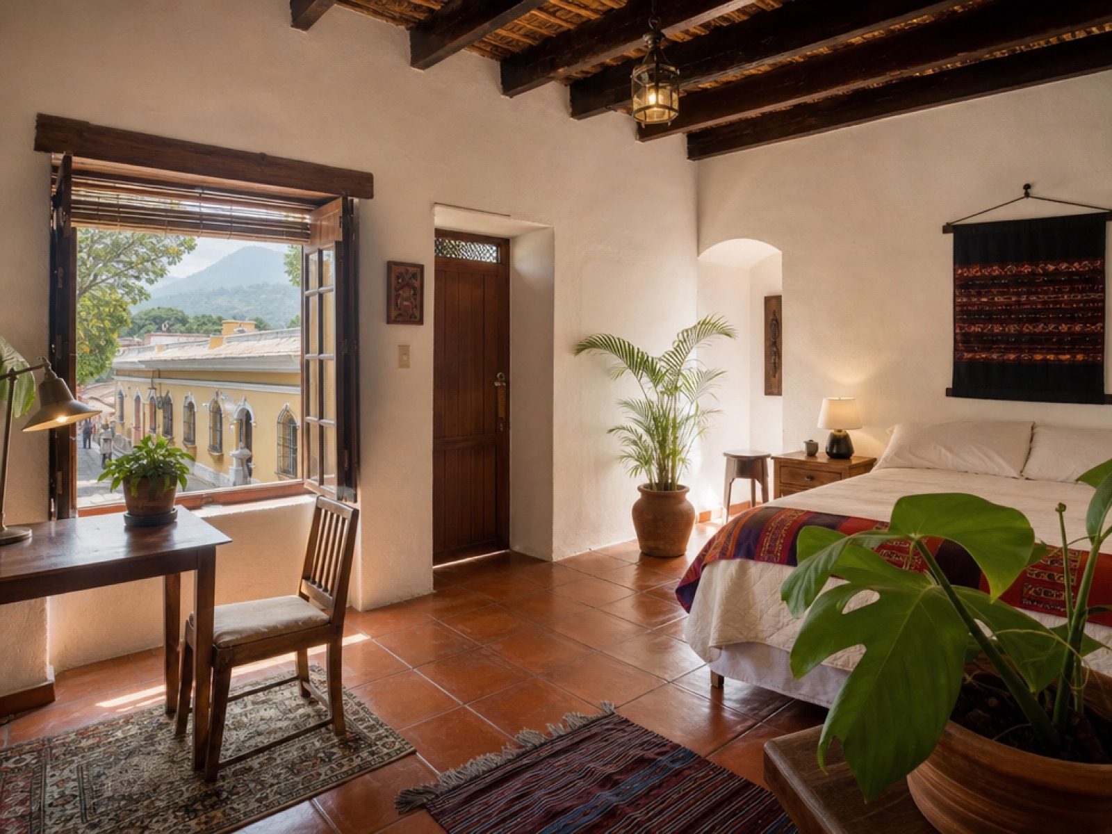 Sunlit furnished bedroom in a colonial Antigua Guatemala rental home, with terracotta tile floor, exposed wooden beams, a desk by an open window, and colorful woven textiles