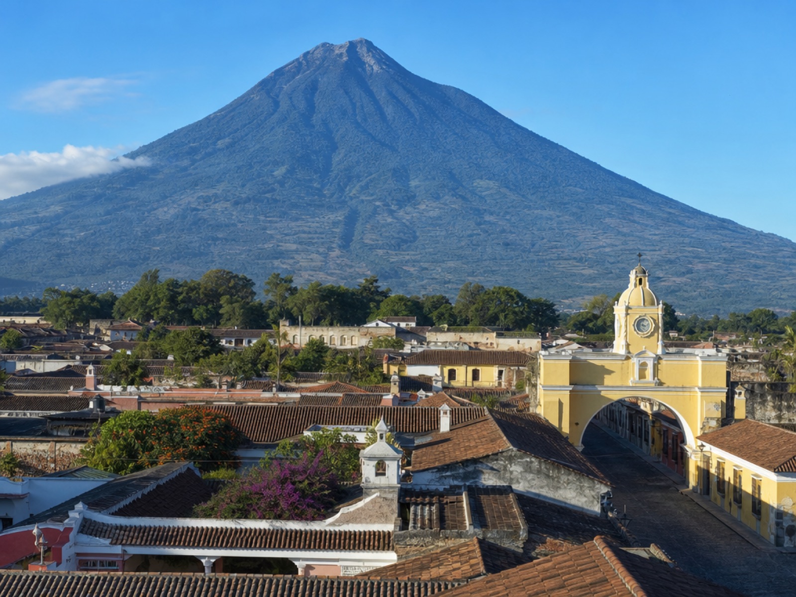 Elevated daylight view of colonial Antigua Guatemala with terracotta rooftops, the yellow Santa Catalina arch and Volcán de Agua behind the town