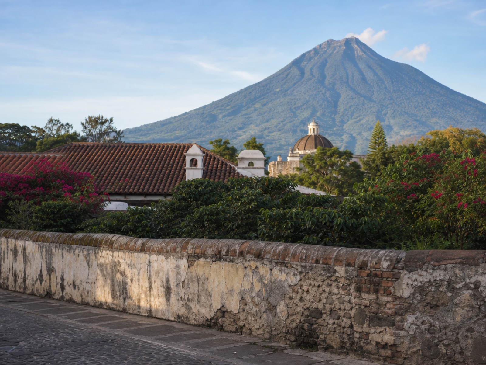 A weathered colonial stone and stucco boundary wall with peeling plaster beside a green garden with pink bougainvillea, terracotta colonial rooftops and a church dome behind, and Volcán de Agua under a clear morning sky in Antigua Guatemala
