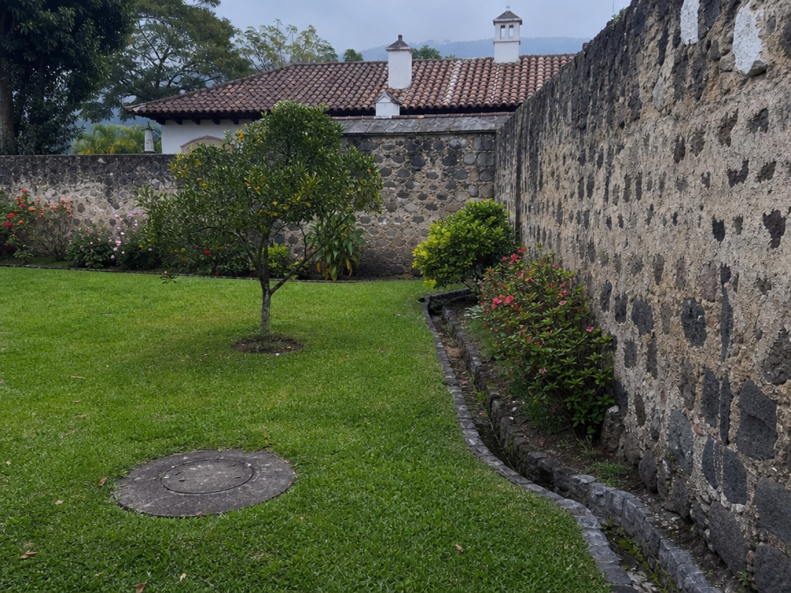 Walled garden of a colonial property near Antigua Guatemala with a round concrete utility cover set in the lawn and a stone drainage channel along a volcanic-stone wall