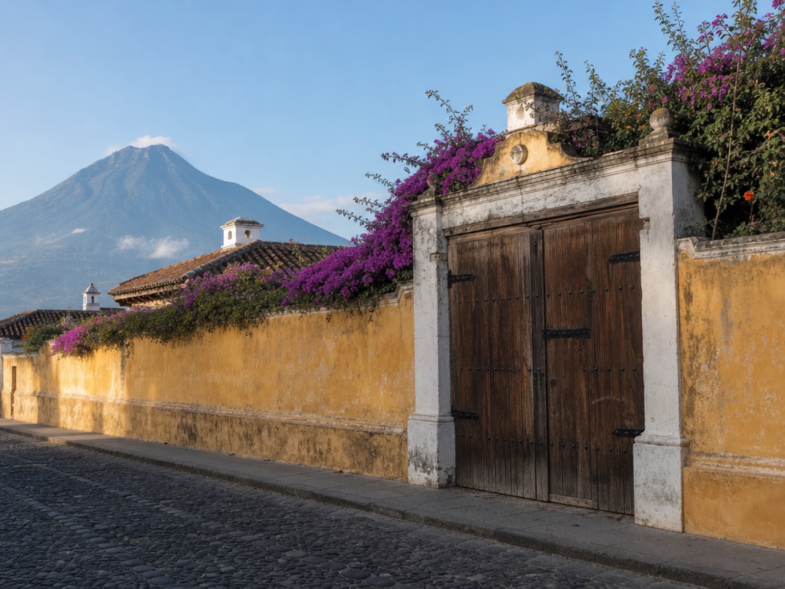 Ochre and white lime-washed perimeter wall along a cobblestone street in Antigua Guatemala with a dark wooden double gate, bougainvillea spilling over the wall and Volcán de Agua rising behind the rooflines